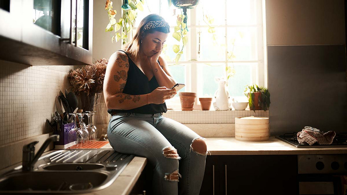 Eine junge Frau sitzt beinebaumelnd auf der Arbeitsfläche der Einbauküche und liest sorgenvoll etwas auf ihrem Smartphone.