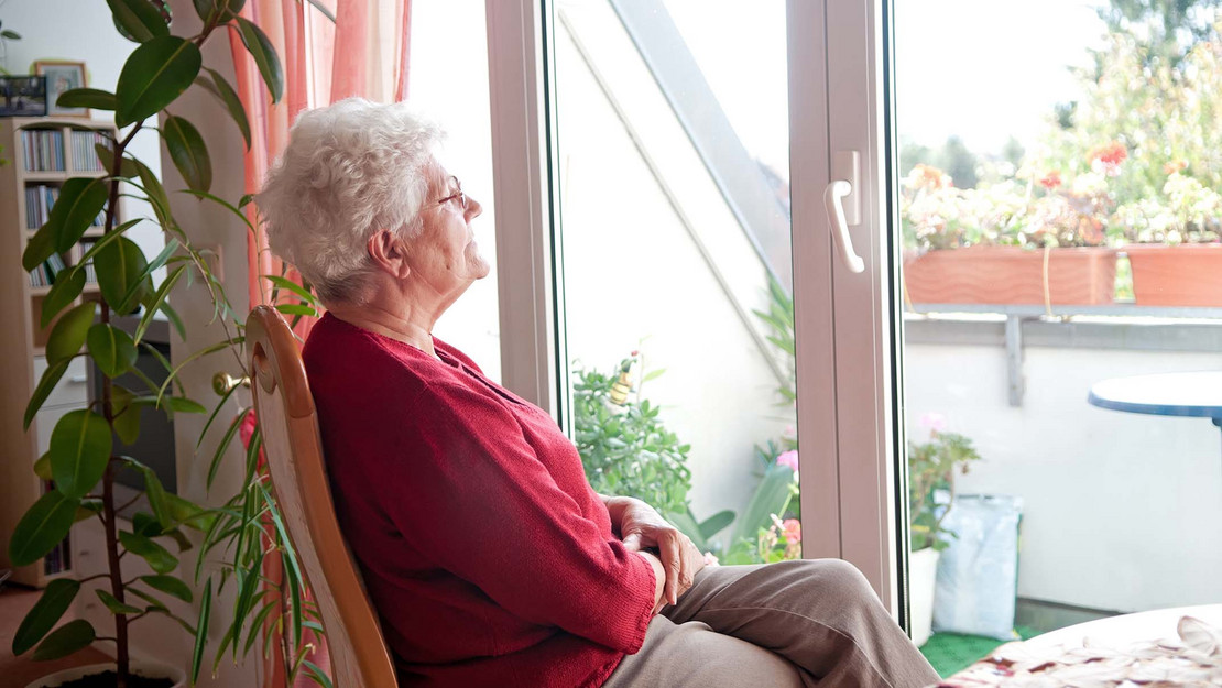 Ältere Frau sitzt in der Wohnung und blickt aus dem Fenster. 