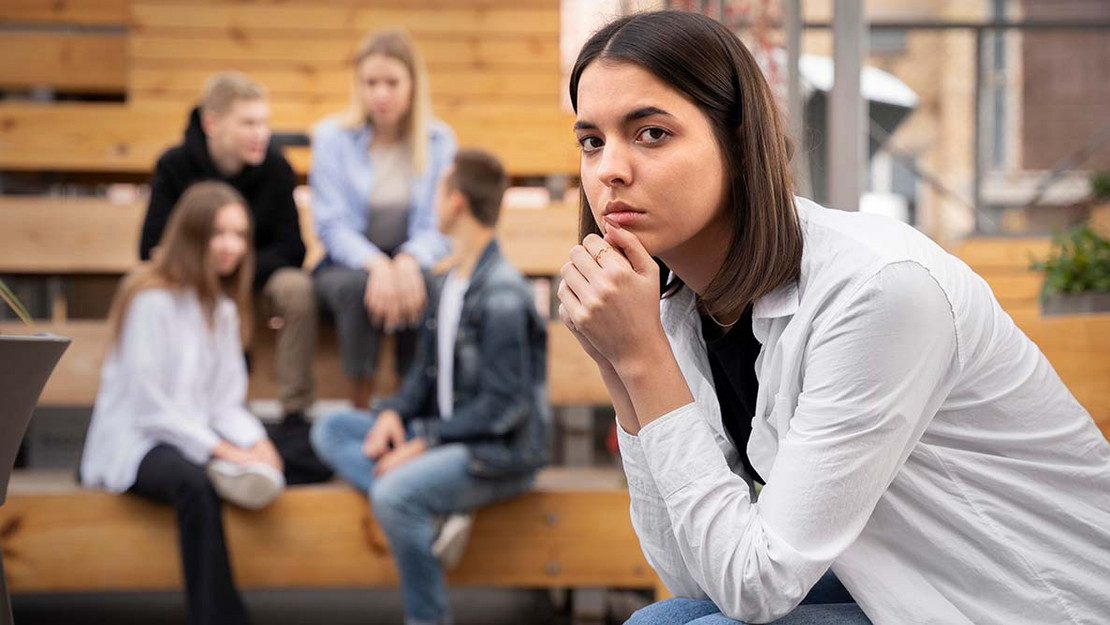 Eine junge Frau blickt besorgt in die Kamera, im Hintergrund reden weitere junge Menschen miteinander. 